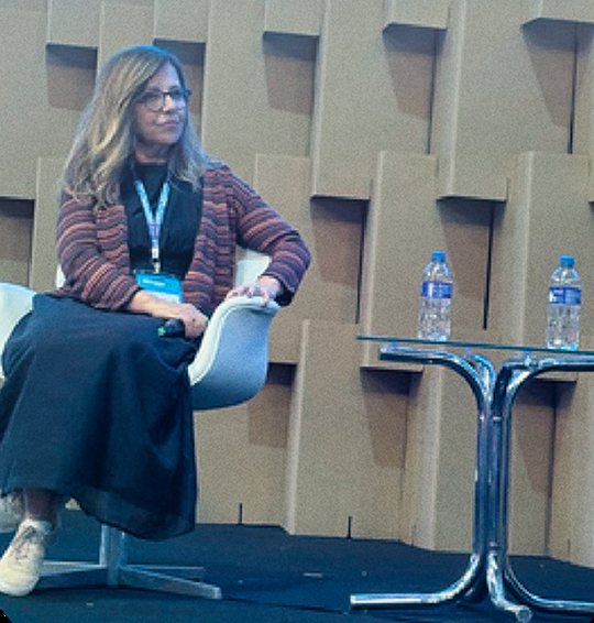 Claudia Fernandes sentada em painel de evento, ouvindo